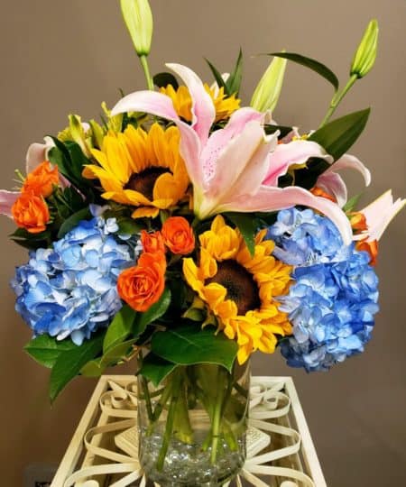 ye-catching star gazer lilies unfold their petals amongst orange spray roses,yellow alstroemeria, and blue hydrangea with lush greens. Clustered in a glass vase, this vibrant bouquet is sure to express your happiest wishes