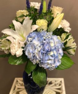 Blue hydrangea and veronica are arranged with beautiful white roses, carnations, lilies and alstroemerias in a blue glass vase.